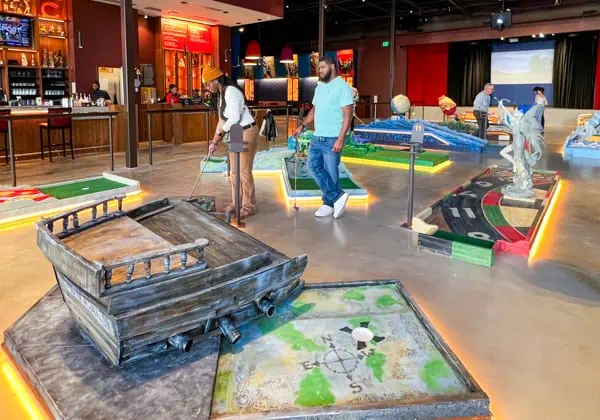 A woman and a man playing indoor miniature golf. The hole features a hand-built wooden pirate's ship.