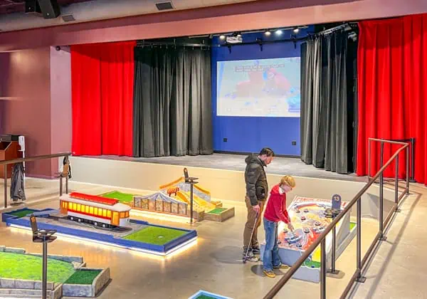 People playing indoor miniature golf. The holes are colorful and bright and lit up. There is a stage in the background with a big movie screen and black and red curtains.