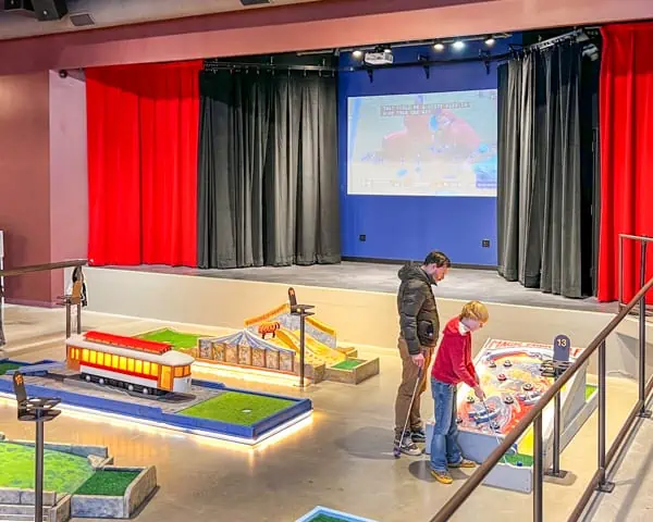People playing indoor miniature golf. The holes are colorful and bright and lit up. There is a stage in the background with a big movie screen and black and red curtains.