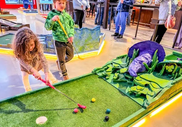 Kids playing mini golf with many different colored golf balls. The hole features a purple hat on rolling green hills