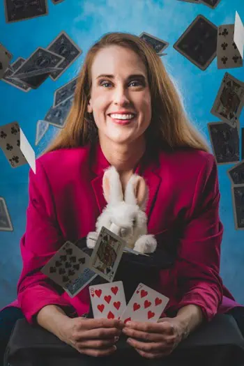 Magician Magic Amanda smiling with a bunny in a top hat and playing cards floating around.